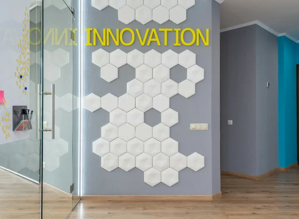 A modern office of a business consulting firm with the word 'INNOVATION' in bold yellow letters on a gray wall, accompanied by a geometric hexagonal white tile design, with a clear glass door to the side and colorful sticky notes on the adjacent glass wall.