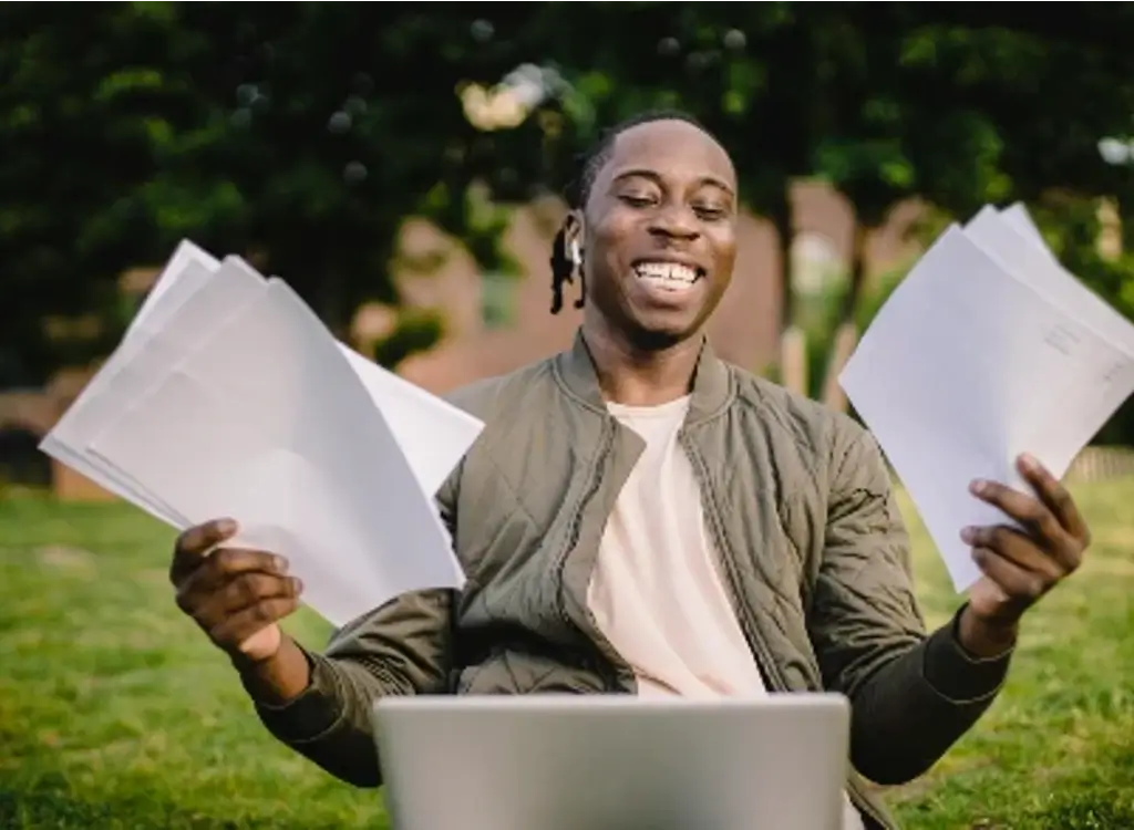 Smiling business owner holding documents and sitting in front of a laptop, symbolizing relief and success after securing emergency business loans.